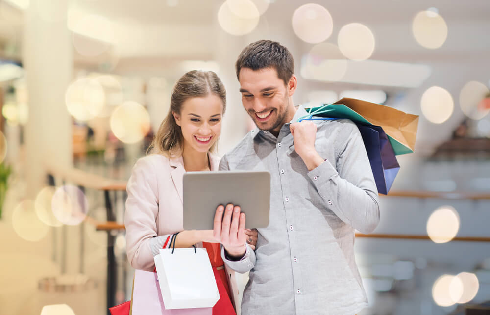 casal usando aplicativo de vendas em tablet segurando sacvolas de compras