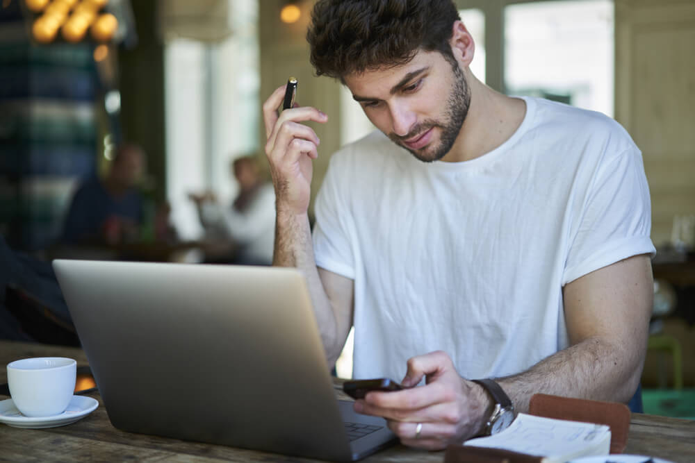 redator pensativo em frente a laptop segurando smartphone com xicara de cafe ao lado ao preparar redaçao publicitaria